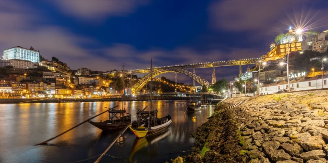 The illuminated Dom Luis I iron bridge over the Douro at dusk, with rabelo boats below